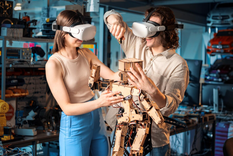 Two people wearing VR headsets work together on a wooden humanoid robot in a tech workshop.